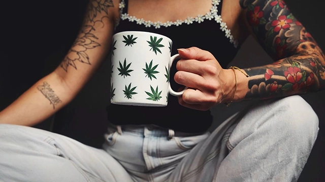 A tattooed individual holding a cannabis leaf-print mug, suggesting the enjoyment of a cannabis shake tea infusion.