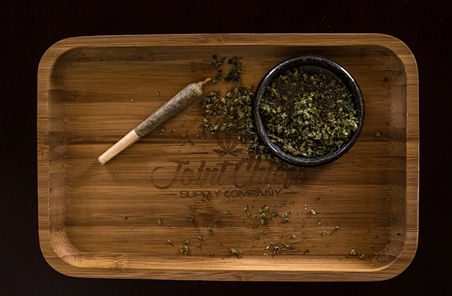 A ready-to-smoke pre-roll on a wooden tray beside a grinder filled with cannabis, showcasing the preparation before smoking.