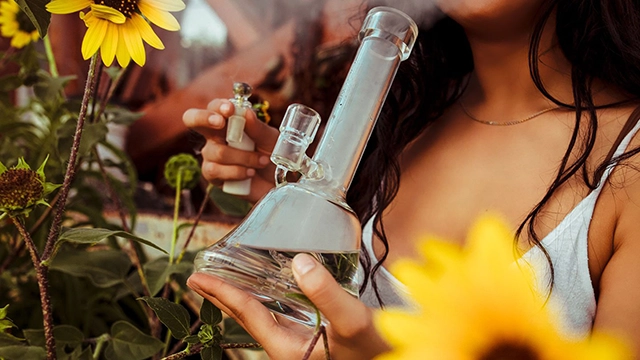 A person lighting a bong surrounded by vibrant flowers, exemplifying one method of consuming cannabis.
